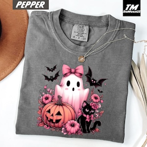 Pode incluir: Uma camiseta cinza Comfort Colors com um gr&aacute;fico tem&aacute;tico de Halloween. O design inclui um fantasma rosa com um la&ccedil;o, uma ab&oacute;bora, um gato preto e morcegos. A camisa tem gola redonda e mangas curtas. O texto "Pepper" est&aacute; no canto superior esquerdo.