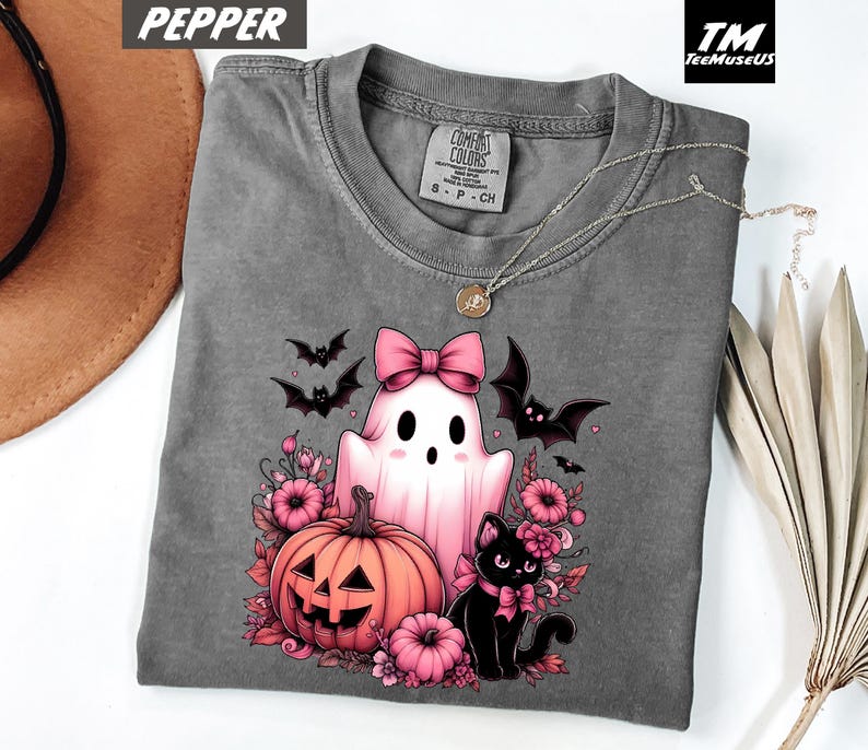 Pode incluir: Uma camiseta cinza Comfort Colors com um gr&aacute;fico tem&aacute;tico de Halloween. O design inclui um fantasma rosa com um la&ccedil;o, uma ab&oacute;bora, um gato preto e morcegos. A camisa tem gola redonda e mangas curtas. O texto "Pepper" est&aacute; no canto superior esquerdo.