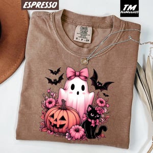 Pode incluir: Camiseta cor espresso com um gr&aacute;fico tem&aacute;tico de Halloween. O design inclui um fantasma rosa com um la&ccedil;o, uma ab&oacute;bora, um gato preto e morcegos. A camisa tem gola redonda e uma etiqueta Comfort Colors.