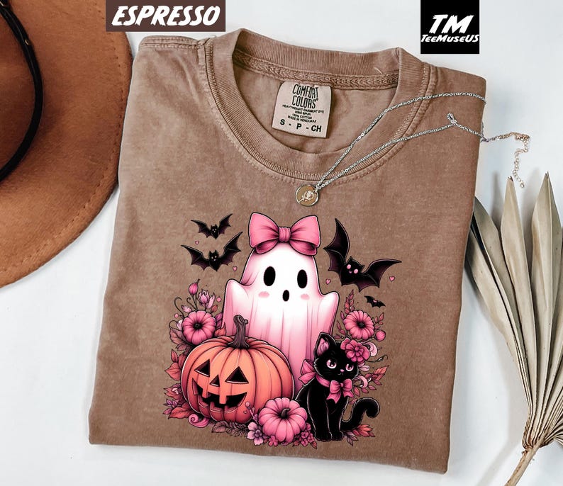 Pode incluir: Camiseta cor espresso com um gr&aacute;fico tem&aacute;tico de Halloween. O design inclui um fantasma rosa com um la&ccedil;o, uma ab&oacute;bora, um gato preto e morcegos. A camisa tem gola redonda e uma etiqueta Comfort Colors.