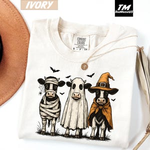 May include: Ivory-colored t-shirt featuring three cows dressed in Halloween costumes. One cow is wrapped as a mummy, another is a ghost, and the third wears a witch hat and cape. Black bats and a spiderweb are also featured.