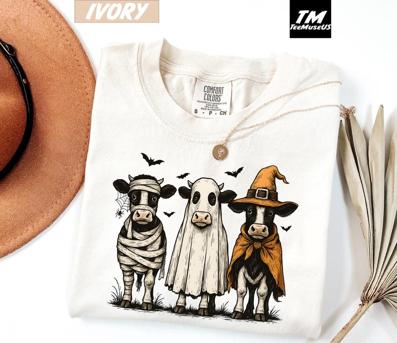 May include: Ivory-colored t-shirt featuring three cows dressed in Halloween costumes. One cow is wrapped as a mummy, another is a ghost, and the third wears a witch hat and cape. Black bats and a spiderweb are also featured.