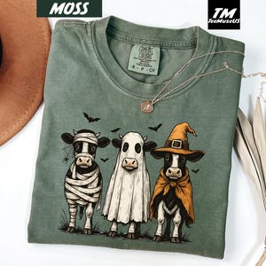 May include: Moss green Comfort Colors t-shirt featuring three cartoon cows dressed in Halloween costumes. One cow is a mummy, one is a ghost, and the third is a witch. The shirt has a crew neck and short sleeves.