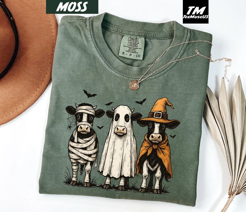 May include: Moss green Comfort Colors t-shirt featuring three cartoon cows dressed in Halloween costumes. One cow is a mummy, one is a ghost, and the third is a witch. The shirt has a crew neck and short sleeves.
