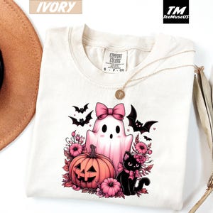 Pode incluir: Camiseta cor marfim com um design de Halloween. O design inclui um fantasma rosa com um la&ccedil;o, uma ab&oacute;bora esculpida, um gato preto e morcegos. A camiseta tem uma etiqueta Comfort Colors e um colar dourado. O logotipo da marca "TeeMuseUS" &eacute; vis&iacute;vel.