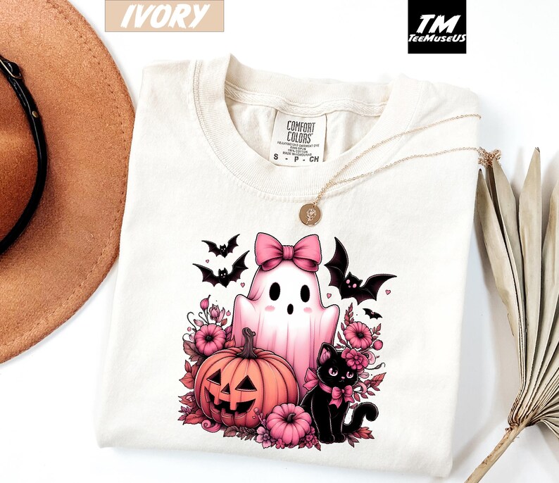 Pode incluir: Camiseta cor marfim com um design de Halloween. O design inclui um fantasma rosa com um la&ccedil;o, uma ab&oacute;bora esculpida, um gato preto e morcegos. A camiseta tem uma etiqueta Comfort Colors e um colar dourado. O logotipo da marca "TeeMuseUS" &eacute; vis&iacute;vel.