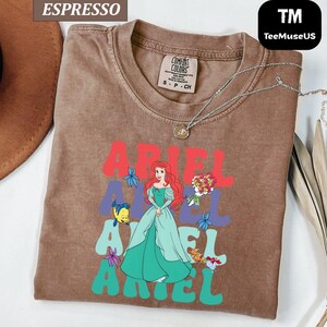 May include: Espresso-colored t-shirt featuring a graphic of Ariel from The Little Mermaid. The design includes the name "ARIEL" in red and teal, with Ariel in a green dress. A silver necklace is draped on the shirt.