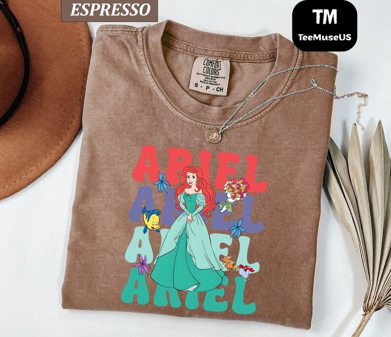 May include: Espresso-colored t-shirt featuring a graphic of Ariel from The Little Mermaid. The design includes the name "ARIEL" in red and teal, with Ariel in a green dress. A silver necklace is draped on the shirt.