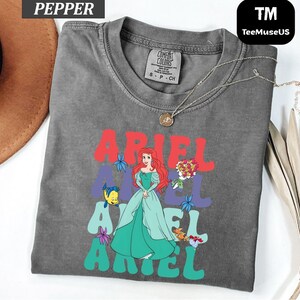 May include: A gray t-shirt featuring a graphic of Ariel from The Little Mermaid. The word "ARIEL" is printed in red and teal, with an illustration of Ariel in a teal dress. The shirt has a crew neck and short sleeves.