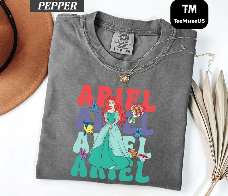 May include: A gray t-shirt featuring a graphic of Ariel from The Little Mermaid. The word "ARIEL" is printed in red and teal, with an illustration of Ariel in a teal dress. The shirt has a crew neck and short sleeves.