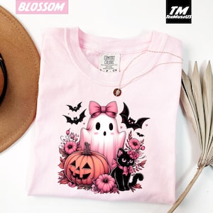 Pode incluir: Camiseta rosa claro com um design de Halloween. O design inclui um fantasma com um la&ccedil;o rosa, uma ab&oacute;bora esculpida, um gato preto e morcegos. A camiseta tem gola redonda e mangas curtas. A marca "Comfort Colors" &eacute; vis&iacute;vel.