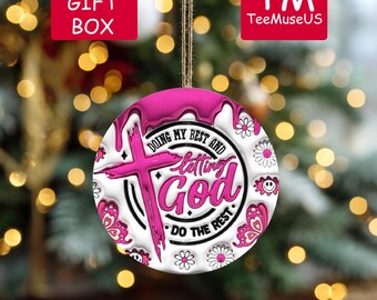 May include: A round Christmas ornament with a pink cross and the words "DOING MY BEST AND letting God DO THE REST." The ornament has a white background with pink floral and butterfly accents. The ornament is hanging from a brown string.