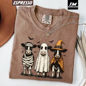 May include: Espresso-colored t-shirt featuring a Halloween-themed graphic of three cows dressed as a mummy, ghost, and witch. The shirt has a crew neck and short sleeves. The brand name "Comfort Colors" is visible on the tag.