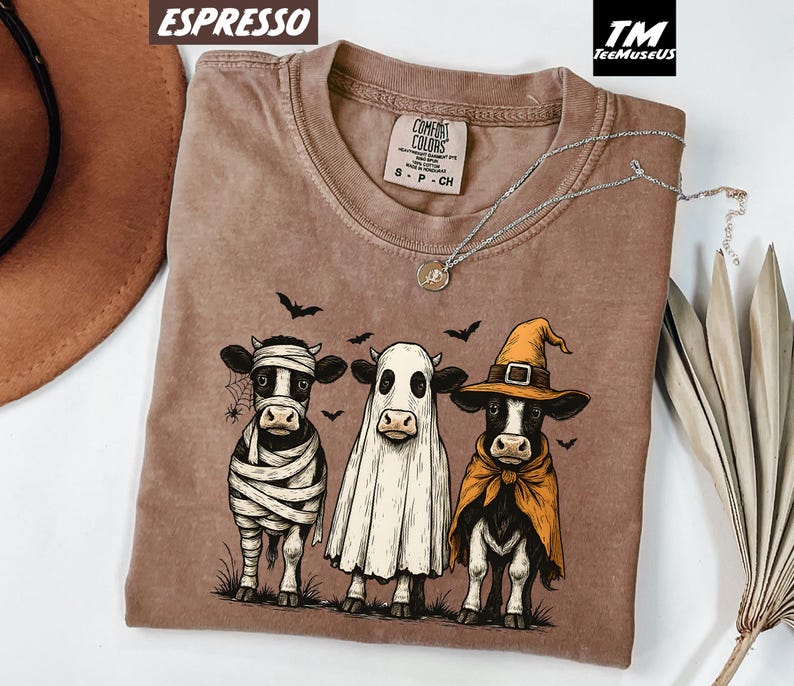 May include: Espresso-colored t-shirt featuring a Halloween-themed graphic of three cows dressed as a mummy, ghost, and witch. The shirt has a crew neck and short sleeves. The brand name "Comfort Colors" is visible on the tag.