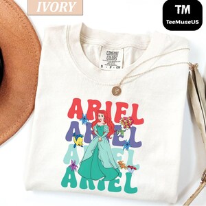 May include: Ivory-colored t-shirt featuring a graphic of Ariel from The Little Mermaid. The design includes the name "ARIEL" in colorful, retro-style lettering, with Ariel's image and other characters. A gold necklace is draped on the shirt.