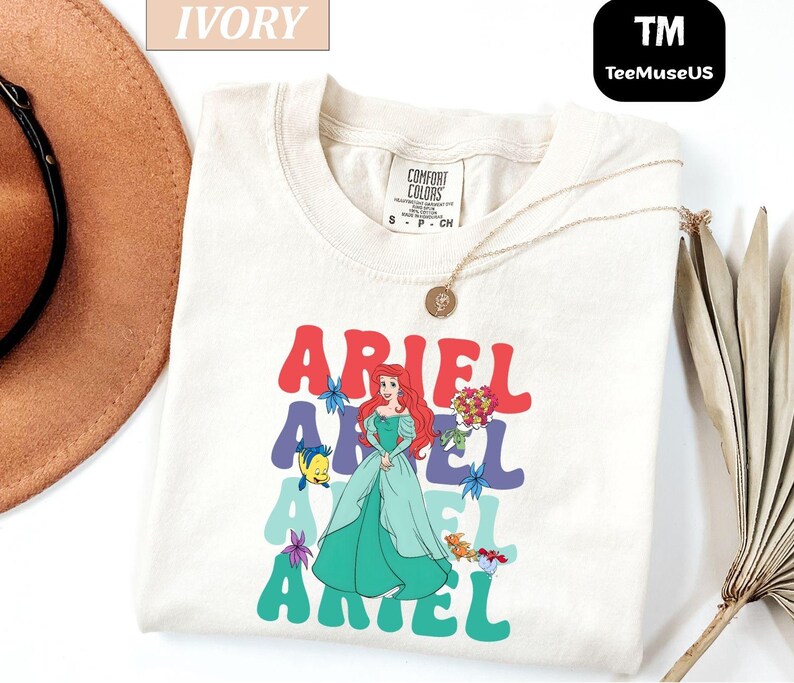 May include: Ivory-colored t-shirt featuring a graphic of Ariel from The Little Mermaid. The design includes the name "ARIEL" in colorful, retro-style lettering, with Ariel's image and other characters. A gold necklace is draped on the shirt.