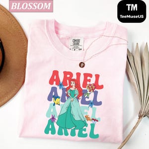 May include: A light pink t-shirt featuring the word "ARIEL" in a retro font, with a cartoon illustration of Ariel from The Little Mermaid. The shirt also includes a necklace and a Comfort Colors tag.
