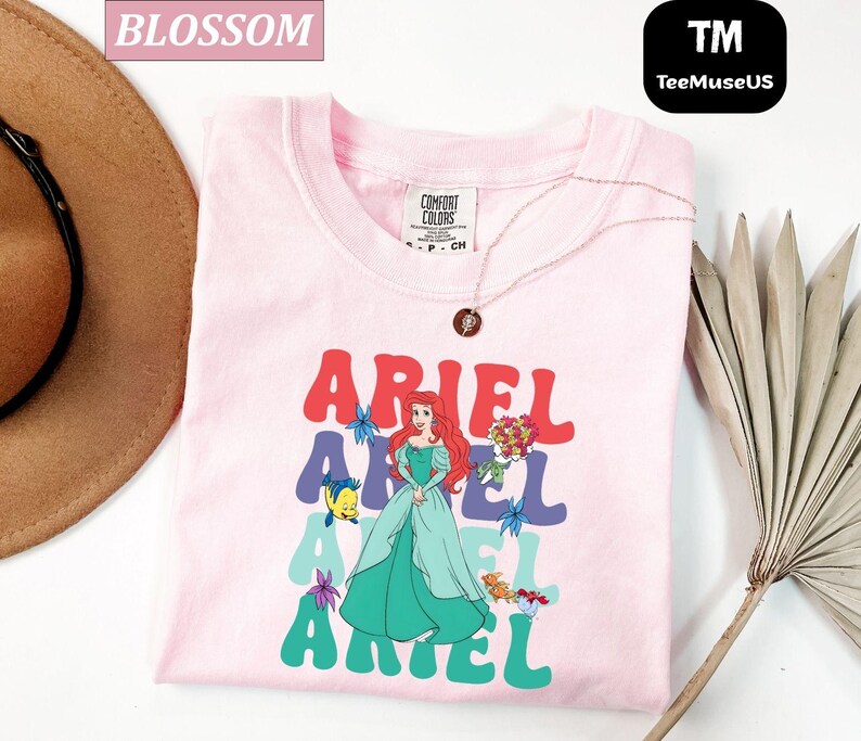 May include: A light pink t-shirt featuring the word "ARIEL" in a retro font, with a cartoon illustration of Ariel from The Little Mermaid. The shirt also includes a necklace and a Comfort Colors tag.