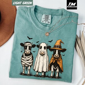 May include: Light green Comfort Colors t-shirt featuring a Halloween-themed graphic of three cows dressed as a mummy, ghost, and witch. The graphic is in black, white, and orange. The shirt has a crew neck and short sleeves. A silver necklace with a pendant is also visible.