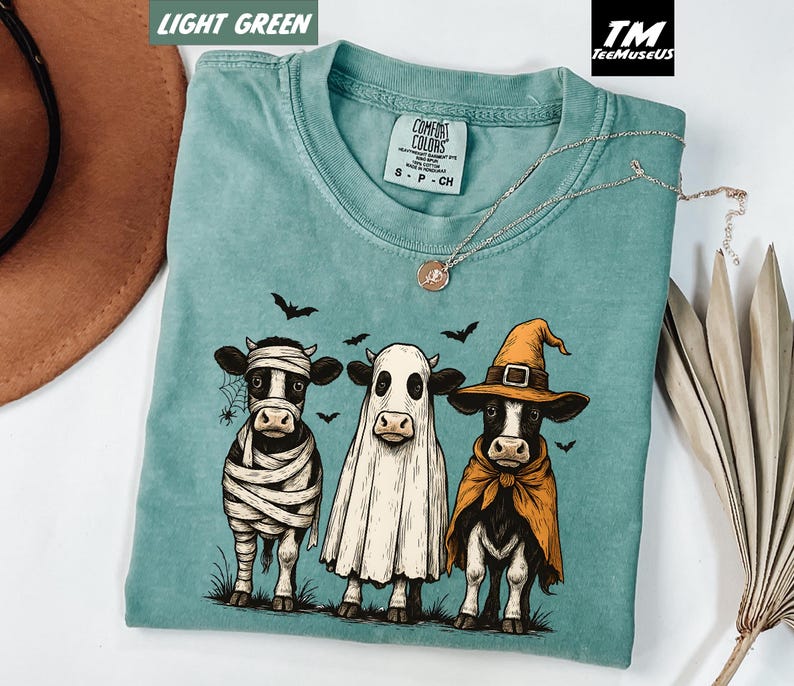 May include: Light green Comfort Colors t-shirt featuring a Halloween-themed graphic of three cows dressed as a mummy, ghost, and witch. The graphic is in black, white, and orange. The shirt has a crew neck and short sleeves. A silver necklace with a pendant is also visible.
