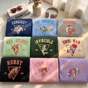 Può includere: Nove felpe piegate in vari colori, ognuna con una diversa grafica e testo di supereroi. Le felpe sono blu navy, azzurro, grigio, verde, verde scuro, crema, nero, rosa e lavanda. Ogni design include il testo "EST. 2021".