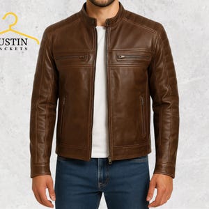 Könnte beinhalten: Braune Lederjacke mit Stehkragen und mehreren Reißverschlusstaschen. Die Jacke hat ein gestepptes Design auf den Schultern und einen Frontreißverschluss. Der Markenname "JUSTIN JACKETS" wird auf einem gelben Kleiderbügel links oben angezeigt.