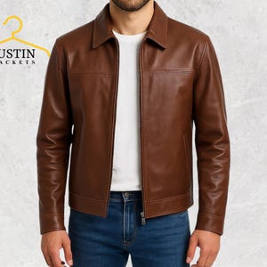 Puede incluir: Chaqueta de cuero marrón con cuello y cierre de cremallera. La chaqueta tiene un diseño clásico con detalles de costura visibles. La imagen también muestra el texto "JUSTIN JACKETS" en amarillo.