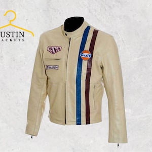 Op de afbeelding: Crèmekleurige leren jas met zwarte kraag en racestrepen. Voorzien van geborduurde patches met "HEUER", "Firestone" en een "Gulf"-logo. De jas heeft een ritssluiting en is van Justin Jackets.