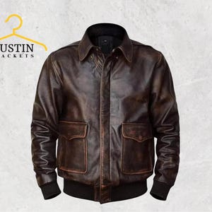 Könnte beinhalten: Braune Lederjacke im Bomber-Stil. Sie hat zwei Vordertaschen, einen Kragen und einen schwarzen gerippten Saum und Bündchen. Der Text "JUSTIN JACKETS" befindet sich über der Jacke.