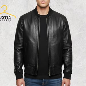 May include: A black leather bomber jacket with a zip-up front and ribbed cuffs and hem. The jacket is worn over a black shirt and blue jeans. The logo "JUSTIN JACKETS" is visible in the upper left corner.