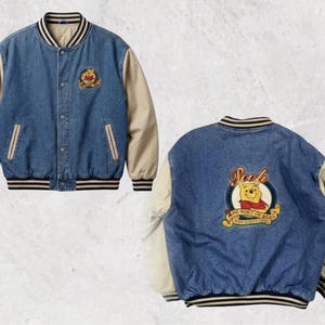 Puede incluir: Chaqueta universitaria de mezclilla azul con mangas beige y detalles a rayas. Presenta parches de Winnie the Pooh en la parte delantera y trasera. Una chaqueta casual con un diseño clásico, ideal para el día a día.