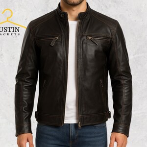 Peut inclure: Veste en cuir marron avec col montant et fermeture éclair. La veste possède deux poches de poitrine zippées et une petite poche sur la manche gauche. Portée sur un t-shirt blanc et un jean bleu. La marque "JUSTIN JACKETS" est visible.