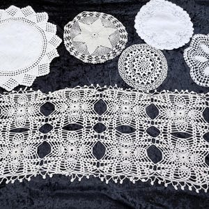 May include: A collection of white crocheted items including one long rectangular table runner with a repeating floral pattern and five circular doilies of varying sizes, each featuring intricate openwork designs and scalloped or picot edges.