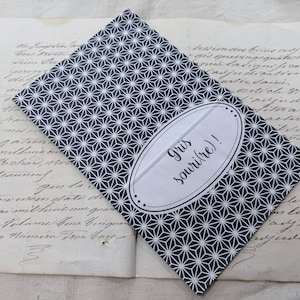 May include: A rectangular notebook featuring a dark blue and white geometric star pattern. An oval white label is centered on the cover, containing the black printed text 'Gris souri(re)!'. The notebook is placed over a sheet of aged, handwritten paper.