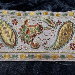 May include: Embroidered trim with a light gray background, featuring paisley and bird designs in gold, green, and rust colors. The trim has a border of small gold and green circles, suitable for various craft projects.