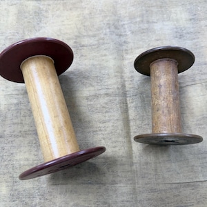 May include: Two cylindrical spools for thread or yarn. The larger spool features a light-colored wooden core with dark maroon circular ends. The smaller spool has a weathered, light brown wooden core with dark brown circular ends.