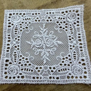 May include: A square white lace doily featuring an intricate needlework pattern with a central floral motif, surrounded by a geometric border of openwork mesh, small circular cutouts, and dense thread stitching along the outer edges.