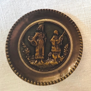 May include: A round, antique bronze button with a detailed relief design. The central image depicts two figures in period clothing against a dark green background. The button's outer edge has a beaded border.