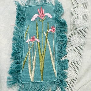 May include: A teal embroidered patch with a fringed edge features pink and white iris flowers with green stems and leaves. The patch is set against a light blue fabric with white lace trim.