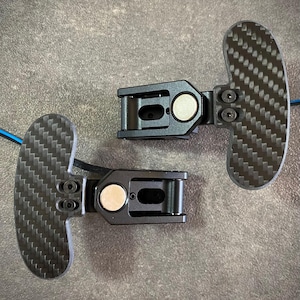 May include: Two black carbon fibre paddle shifters with a textured surface. Each shifter has a black mounting bracket with a silver-coloured circular magnet. The paddles are connected by blue wires.