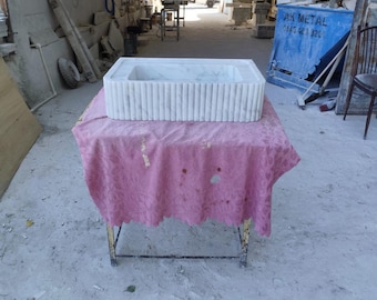 Pode incluir: Uma pia retangular de mármore branco com detalhes de ranhuras verticais. A pia é exibida em uma pequena mesa de metal coberta com uma toalha de mesa rosa. O fundo mostra uma oficina com várias ferramentas e materiais.