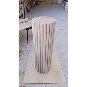 Può includere: Un piedistallo alto, cilindrico, in pietra beige con scanalature verticali. La parte superiore del piedistallo ha un bordo smerlato. Il piedistallo è appoggiato su una tavola di legno chiaro. Lo sfondo è un pavimento in cemento.