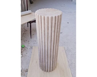 May include: A tall, cylindrical, beige stone pedestal with vertical fluting. The top of the pedestal has a scalloped edge. The pedestal is standing on a light-colored wooden board. The background is a concrete floor.