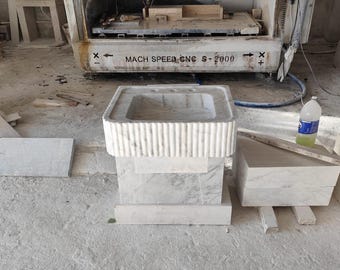 Pode incluir: Uma pia de mármore branco com uma bacia retangular e uma borda canelada. A pia fica em cima de uma base de pedestal, com uma máquina CNC ao fundo. O texto "MACH SPEED CNC S-2000" é visível na máquina.