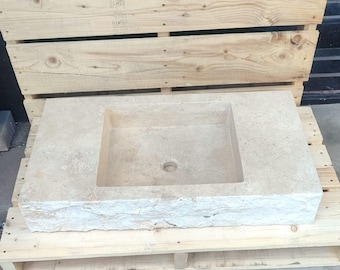 Op de afbeelding: Een rechthoekige, beige stenen wastafel met een vierkant bekken en een centraal afvoergat. De wastafel heeft een ruwe, getextureerde rand en staat op een houten pallet. De wastafel is ontworpen voor gebruik in de badkamer.