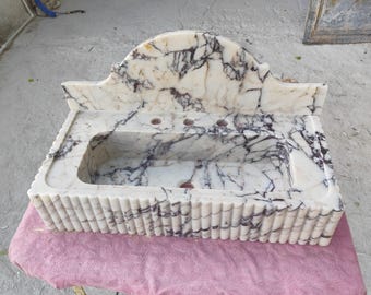 May include: A white marble sink with dark gray veining. The sink has a rectangular basin with three faucet holes and a decorative, fluted edge. The back of the sink has a curved, arched design.