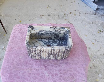May include: A rectangular marble sink with a white and dark gray pattern. The sink has a textured, ridged exterior and sits on a pink floral patterned cloth. The sink is a unique bathroom fixture.