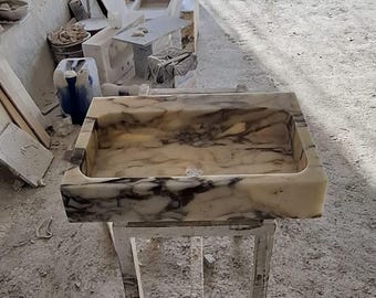 May include: A rectangular marble sink with a cream and gray marbled pattern. The sink is set on a white metal stand. Other marble fixtures are visible in the background.