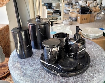 May include: A black marble bathroom set with white veining. The set includes a soap dispenser, a toothbrush holder, a covered jar, a small dish, and a tray. The items are arranged on a round, textured surface.