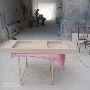 Peut inclure: Un évier de salle de bain rectangulaire en pierre de couleur claire avec deux vasques et une étagère intégrée. L'évier est soutenu par une table métallique recouverte d'un tissu rose. L'évier est dans un atelier.